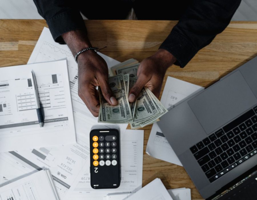Person counting US dollars, using a calculator and laptop, with financial documents on a wooden desk.